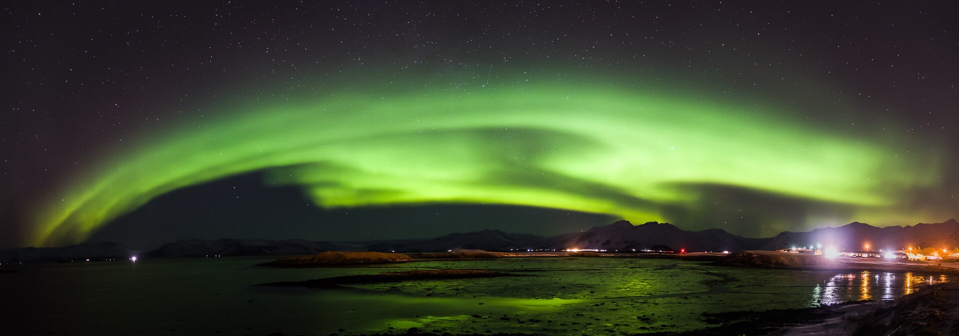 Wide aurora arch over the bay and town lights at Höfn